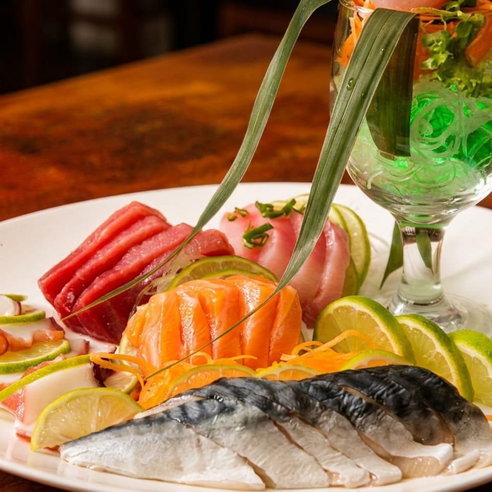 Colorful sashimi platter with salmon, tuna, mackerel, octopus, lime wedges, vegetables, and a green garnished cocktail on a wooden table.